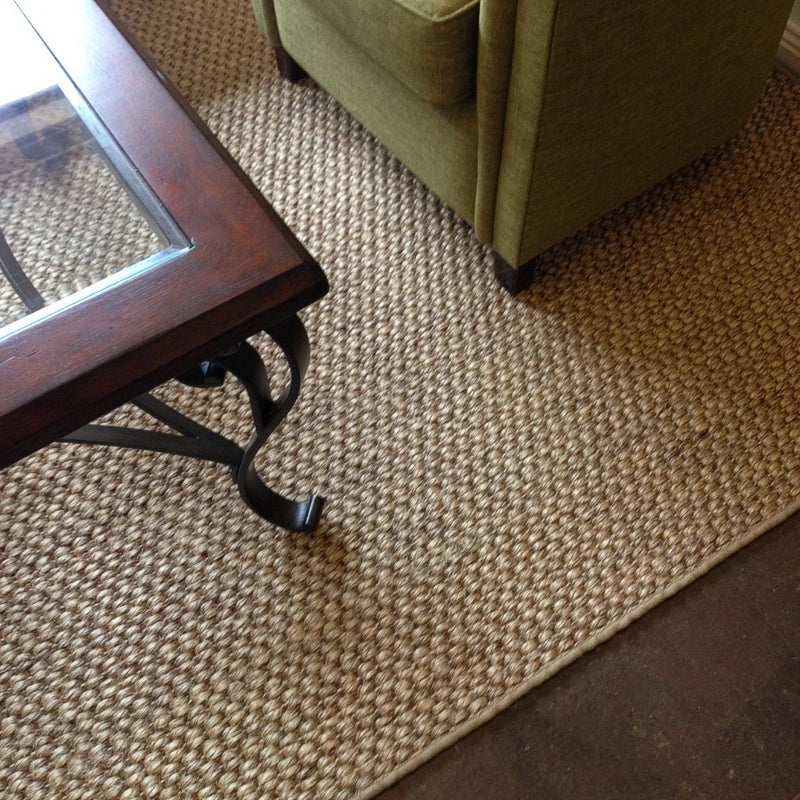 A green armchair and glass-top table rest atop a richly textured, rustic sisal rug on warm brown flooring in a refined living space.