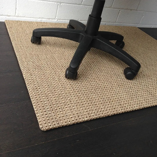 A black office chair sits on a natural sisal chair pad in Autumn Slate, atop dark timber flooring by a white brick wall.