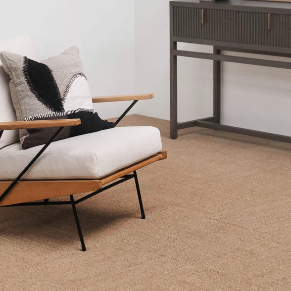 A modern armchair with a white cushion and patterned pillow sits beside a grey console on natural sisal tiles in a styled interior.