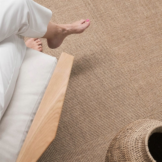 Barefoot individual in white pants sits on natural sisal tiles, beside a woven basket and wooden chair in a serene, styled interior.