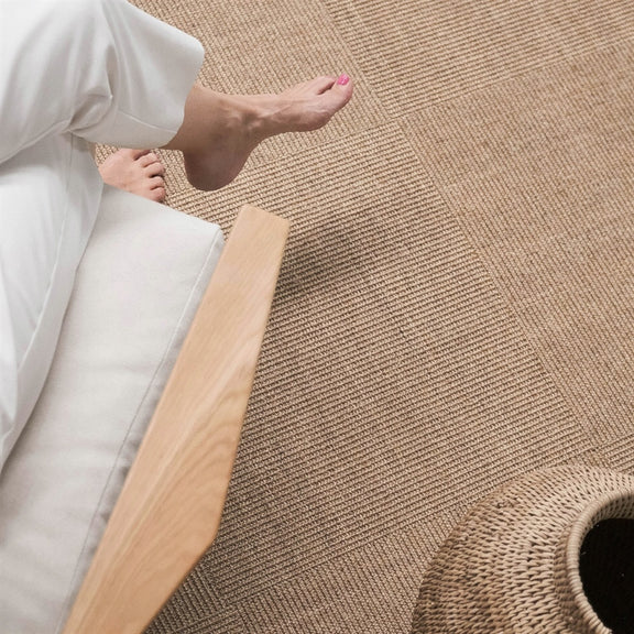 Barefoot individual in white pants sits on natural sisal tiles, beside a woven basket and wooden chair in a serene, styled interior.