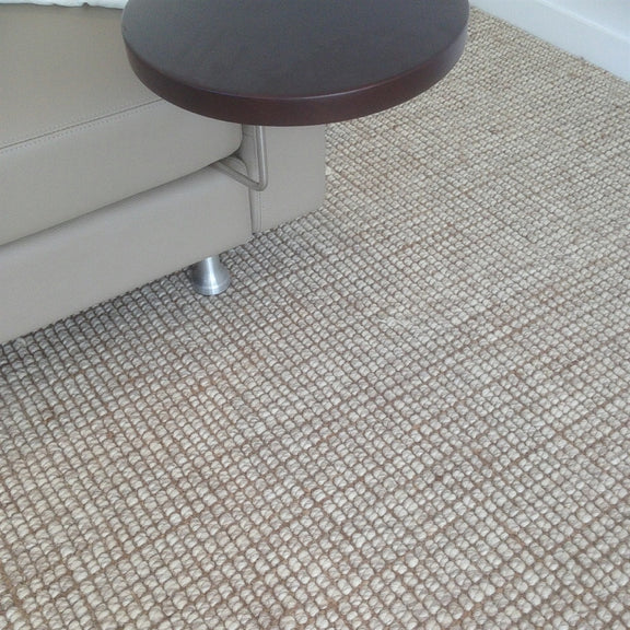 A round wooden side table sits beside a beige sofa on a Snowgum wool and jute rug in a sunlit, elegantly styled living room.