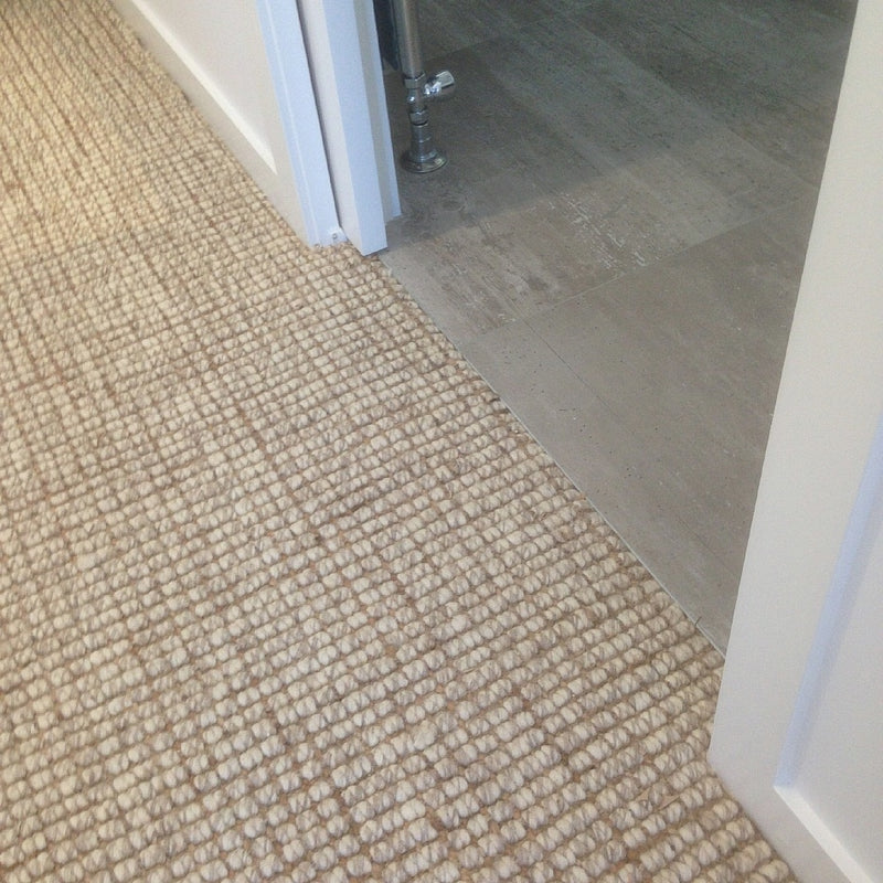 Natural wool and jute flooring transitions seamlessly to pale gray tile at a doorway with crisp white trim in a bright interior.