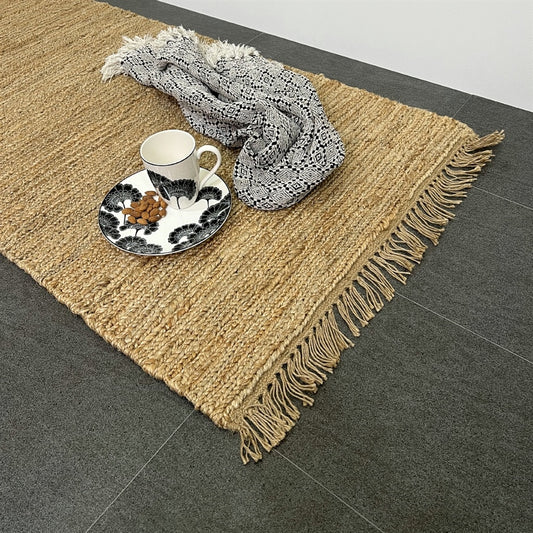 A ceramic cup and snack plate rest on a monochrome tray beside a folded scarf atop a textured natural jute runner in a styled interior.
