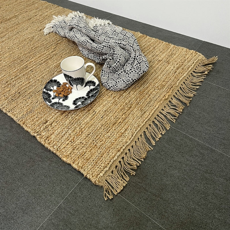 A ceramic cup and snack plate rest on a monochrome tray beside a folded scarf atop a textured natural jute runner in a styled interior.