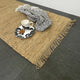 A cup and saucer with snacks sit atop a terra firma jute runner layered over patterned fabric on dark tiled flooring in a styled setting.