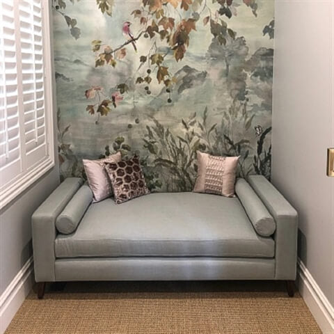 A pale blue sofa with cushions sits on finely crafted natural fibre flooring, beside a mural and sunlit window with white shutters.