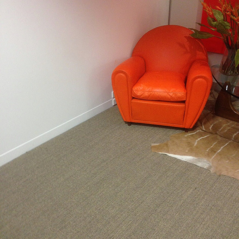 A vibrant orange armchair sits beside a glass table topped with orange flowers on fine ribbed granite sisal in a styled interior.