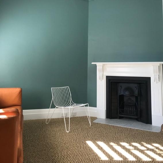 Sunlit room with teal walls, a white fireplace, wire chair and elegant natural fibre flooring in a thoughtfully styled interior.