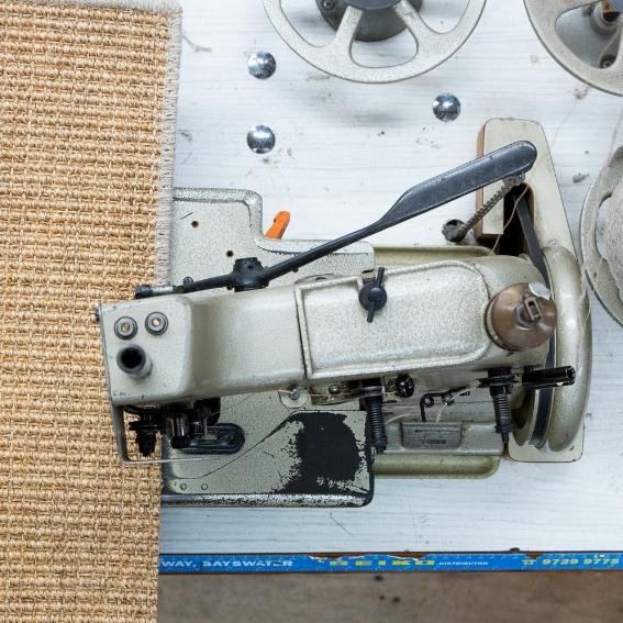 Close-up of an industrial sewing machine expertly finishing the edge of a textured natural fibre rug, highlighting skilled craftsmanship.