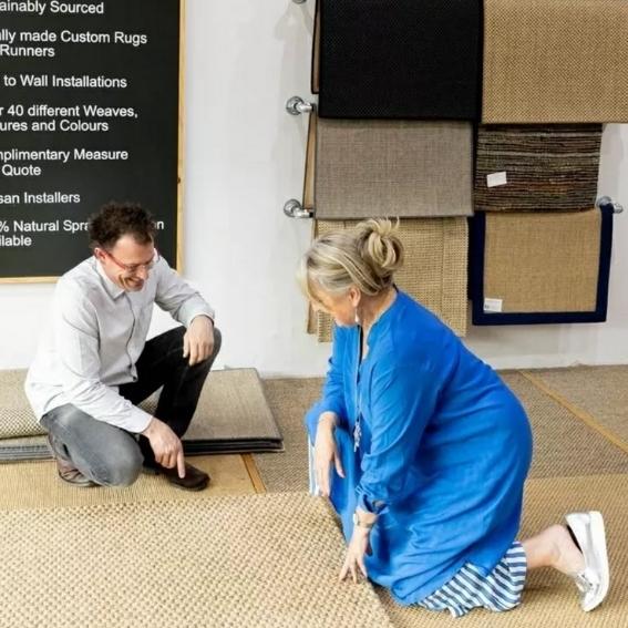 A man and woman thoughtfully compare natural fibre rug swatches on the floor of an elegant, light-filled flooring showroom.
