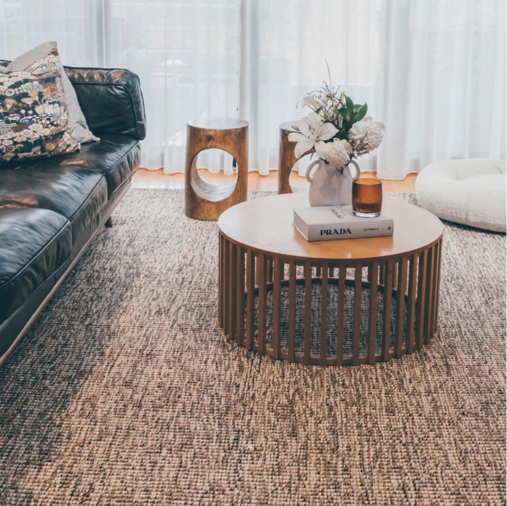 A round wooden coffee table with books and flowers rests on premium natural fibre flooring beside a black sofa in a sunlit lounge.
