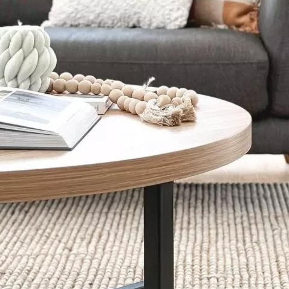 A round wooden coffee table with an open book and decor, set on a natural fibre rug before a dark sofa in a styled living space.