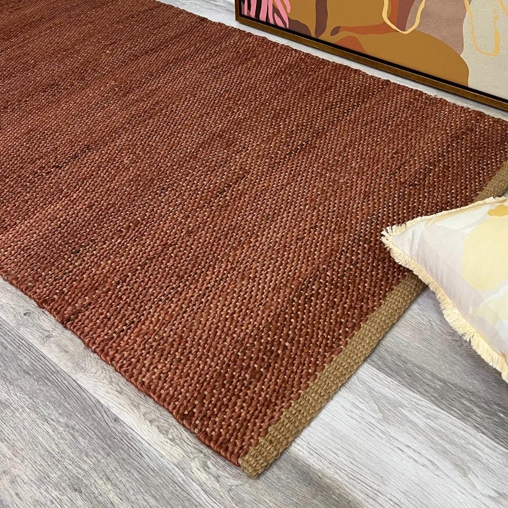 A handwoven brown natural fibre rug rests on pale timber flooring, styled with a cream cushion and vibrant contemporary artwork.