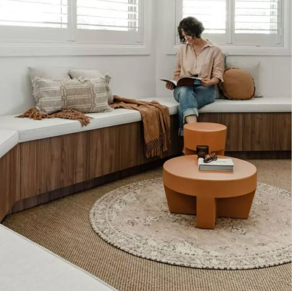 A woman sits reading on a bench in a refined living room with sculpted round tables and a handwoven natural fibre rug underfoot.