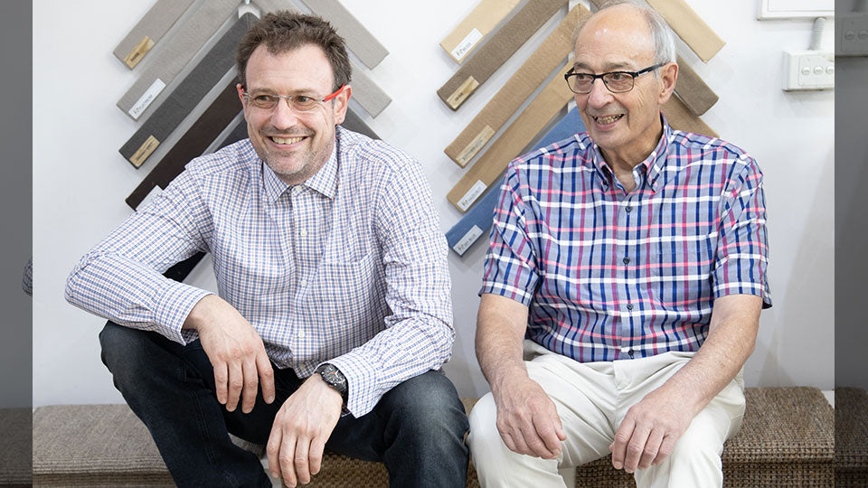 Two men in plaid shirts smile, seated before displays of high-quality natural fibre rugs and elegant premium flooring samples.