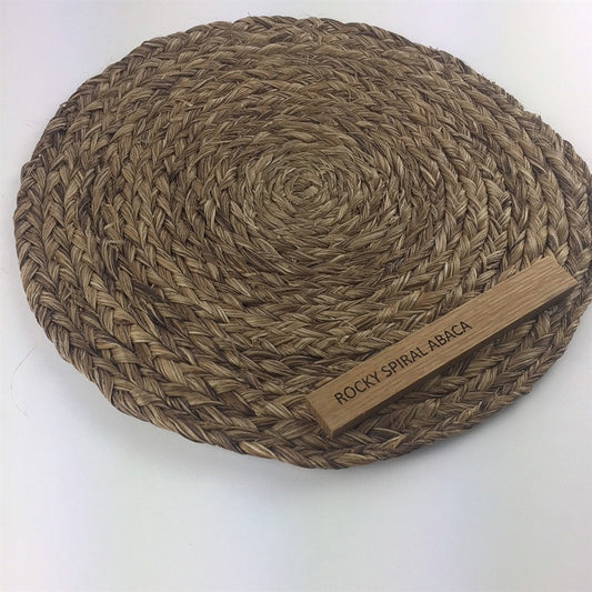 Round placemat with intricately spiraled abaca weave, natural tones, and a small wooden tag detail on the edge, set on a tabletop.